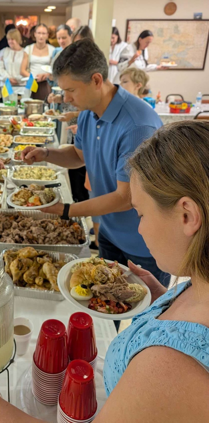 The potluck table in the parish hall for Ukraine's Independence Day: trays of food down a long table with small Ukrainian flags, parishioners filling their plates